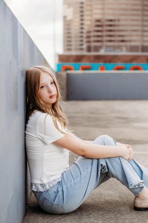 Young blonde woman in white top and jeans sitting against concrete wall on urban rooftop.