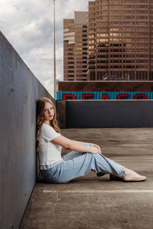 Young woman in white tee and jeans sits against concrete wall on rooftop with city skyline backdrop.