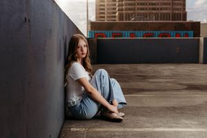 Young woman in white tee and jeans sitting against a concrete wall on a rooftop parking garage with city skyline.