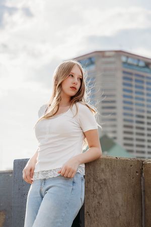 Blonde woman in white lace-trim tee and jeans posing on rooftop with city skyline behind her.
