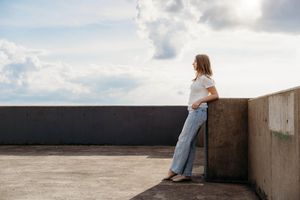Young woman in white top and blue jeans leaning against concrete wall on rooftop under cloudy sky.