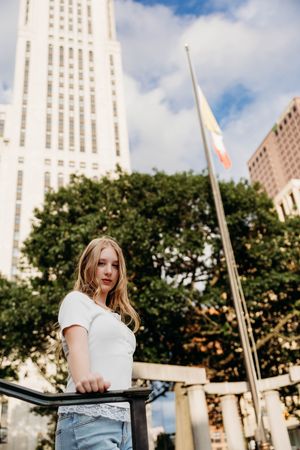 Young blonde woman in white top and jeans poses by a railing with city buildings and a flag in the background.