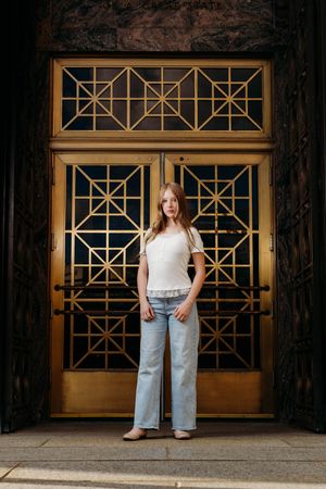 Young woman in white top and light jeans stands before ornate gold Art Deco doors in an urban setting.