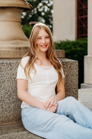 Smiling teenage girl with long blonde hair sitting on stone steps wearing a white top and light blue jeans outdoors.