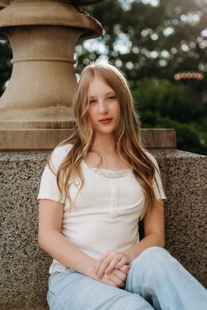 Young woman with long blonde hair sits by a stone fountain, wearing a white top and jeans, gazing at the camera.