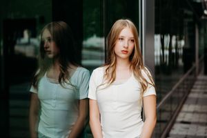 Young blonde woman in white t-shirt stands beside reflective glass wall outdoors, her reflection visible behind her.