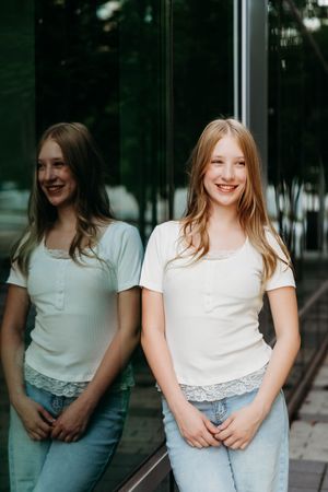 Smiling teenage girl in white t-shirt and jeans standing by reflective glass wall outdoors.