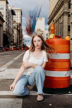 Young girl in white tee and jeans sits beside orange construction barrel on urban city street.