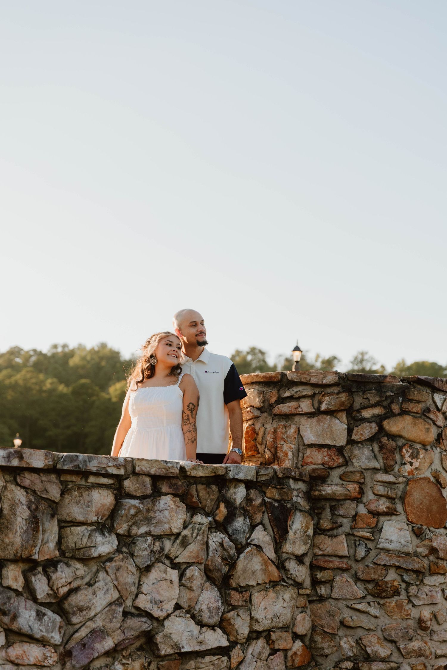 A tattooed couple stands together on a stone wall outdoors, with trees and a clear sky in the background.