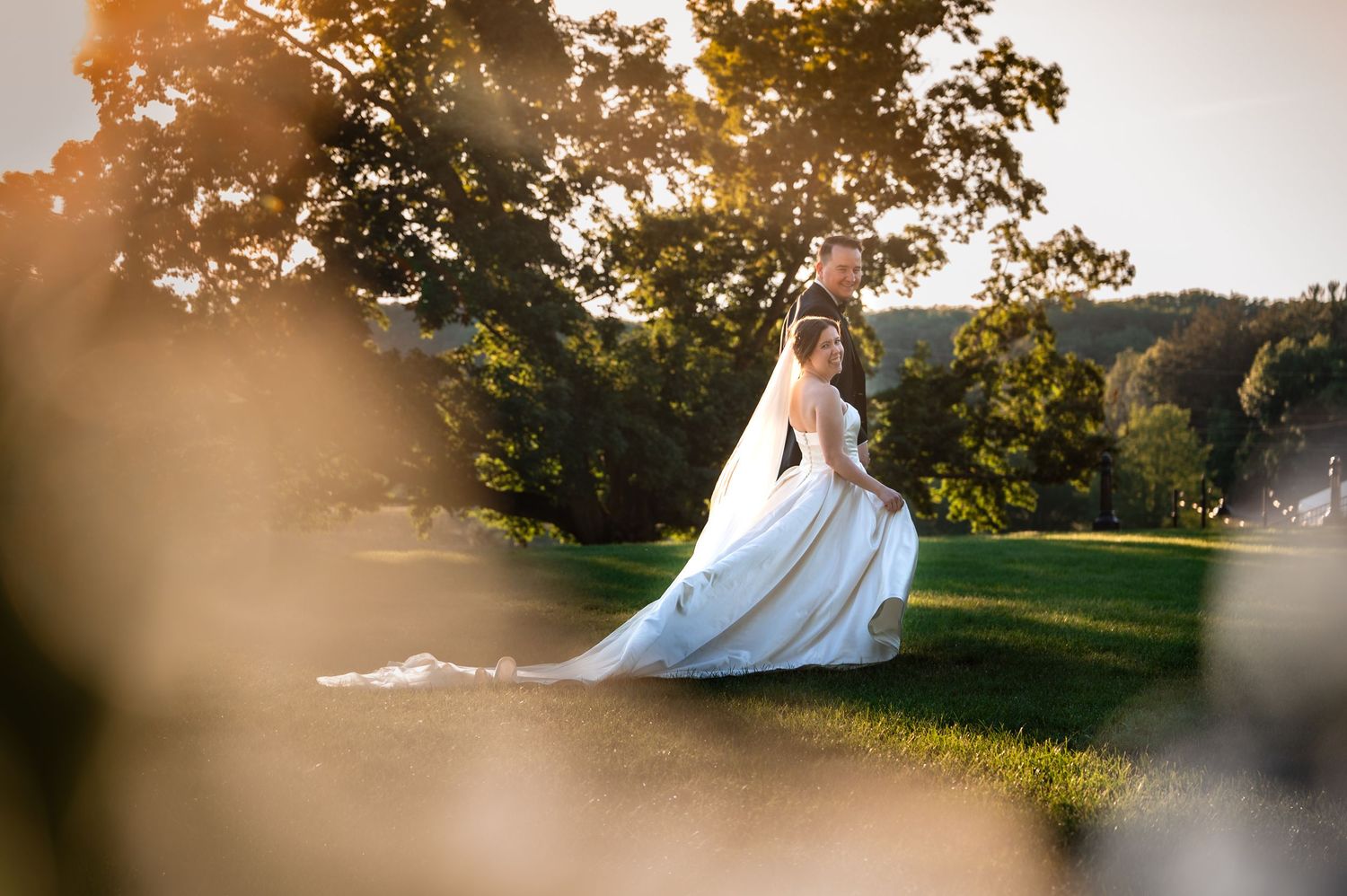 Bride and groom share a romantic moment on a sunlit lawn with golden hour light filtering through lush green trees.
