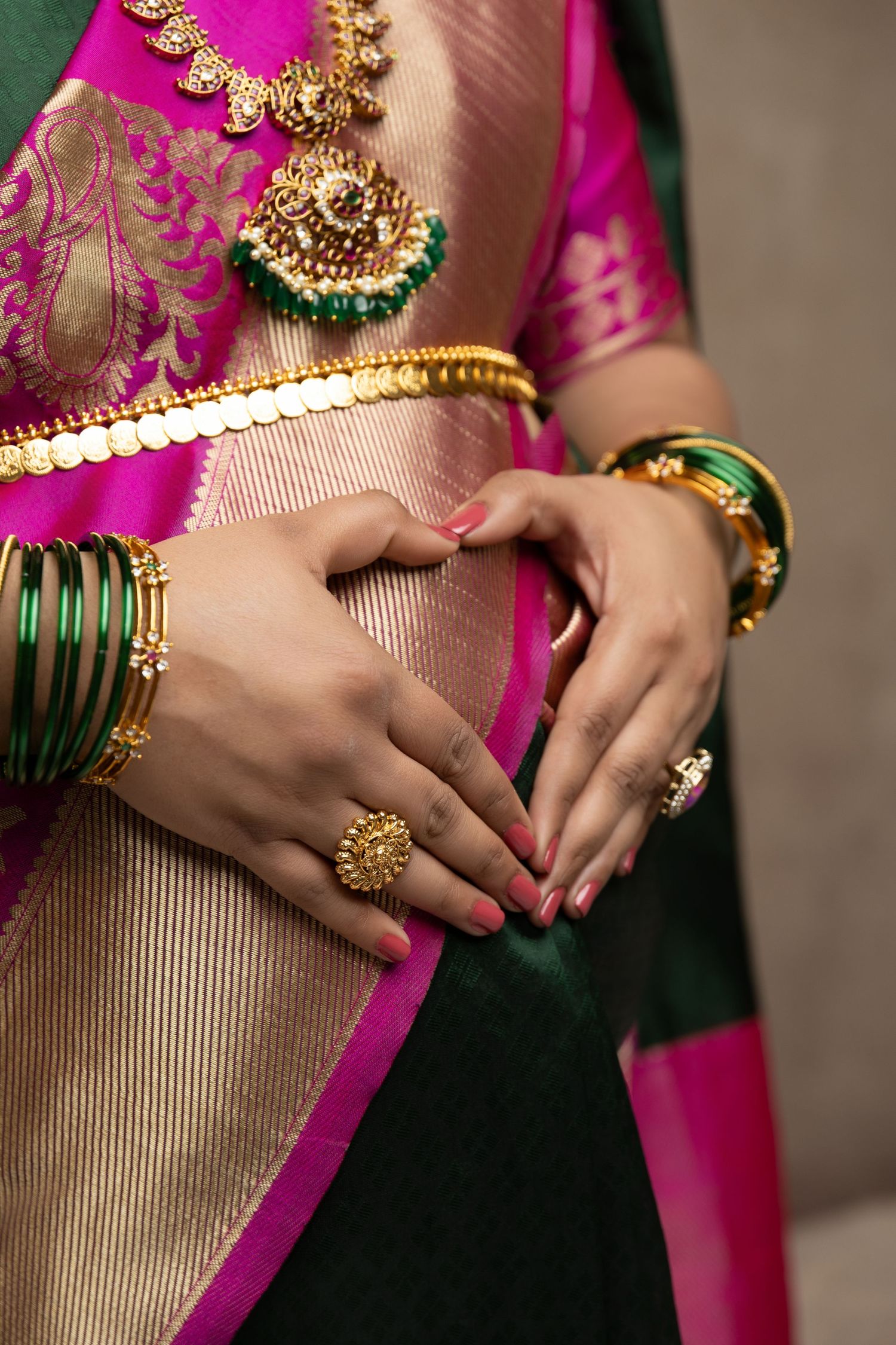 Woman in pink and gold saree with green bangles, gold rings, and traditional Indian jewelry posing elegantly.