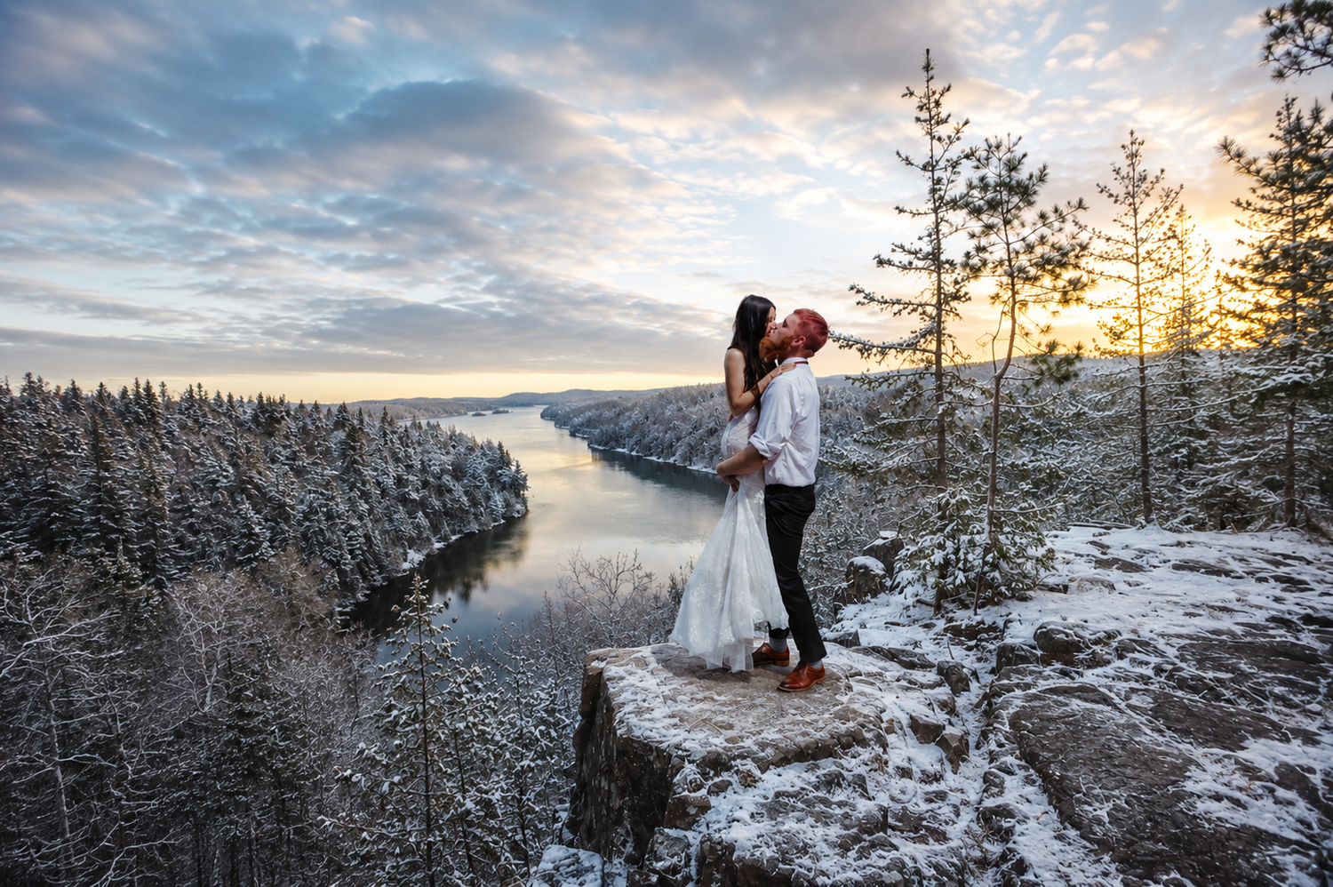 Couple embracing on snowy cliff edge at sunset, surrounded by snow-covered pine trees and a winding river below.