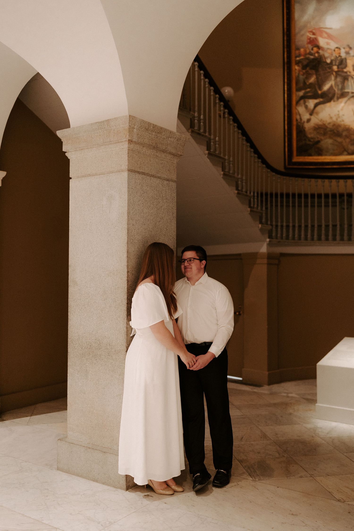 A couple in white outfits share a romantic forehead touch in an elegant hallway with classic artwork.