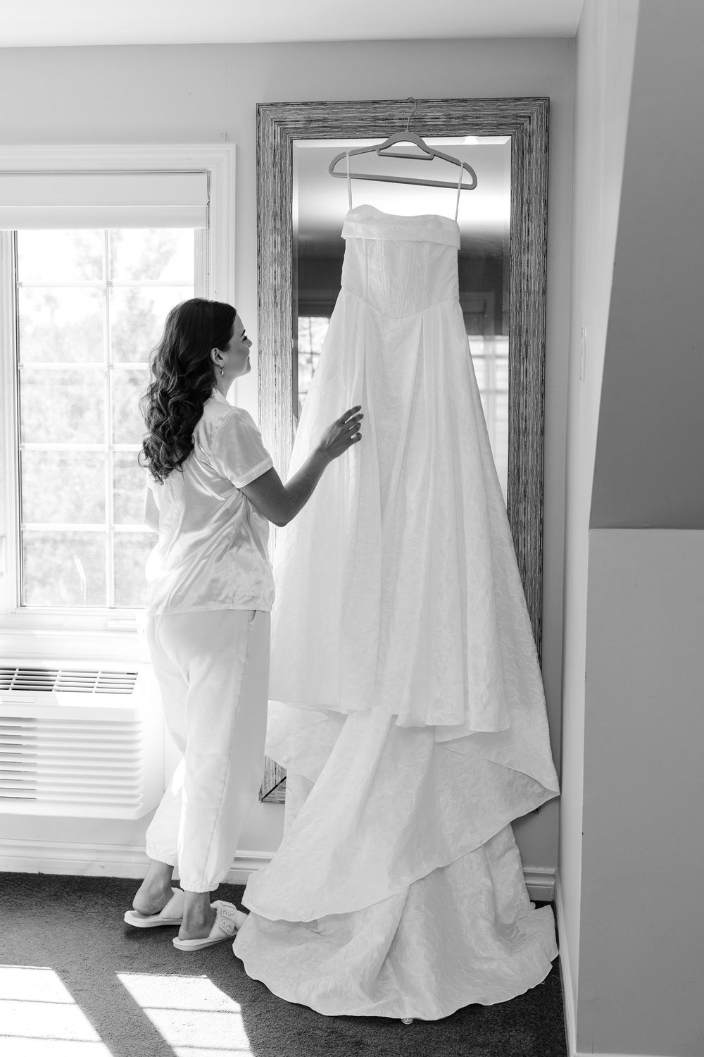 Bride in robe admiring her white wedding gown hanging on a mirror in a sunlit room, black and white photo.