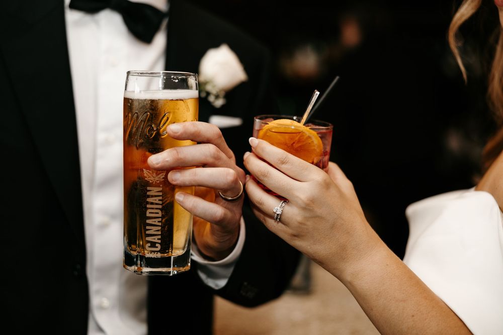 Couple toasting with cocktails at a wedding reception, holding a Canadian whiskey highball and an Aperol spritz.
