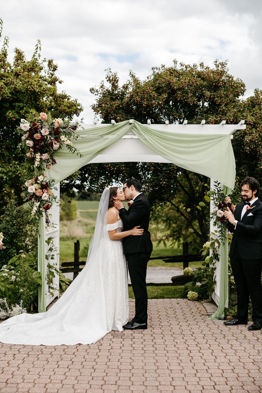 Bride and groom share first kiss under floral arch with sage green draping at outdoor wedding ceremony.