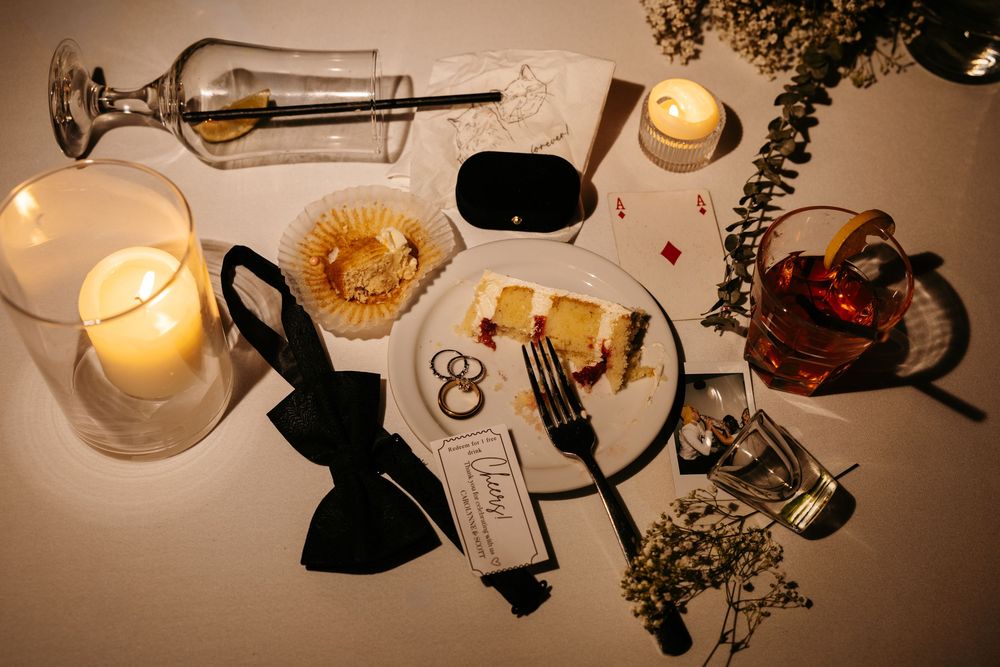 Elegant wedding table setting with candles, bow tie, rings, cake slice, and florals in warm candlelight.