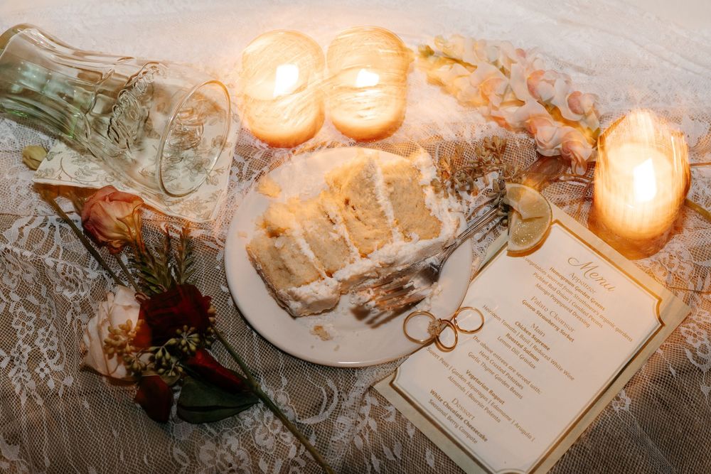 Elegant wedding cake slice on a plate surrounded by candles, flowers, and a gold-trimmed menu card on lace.