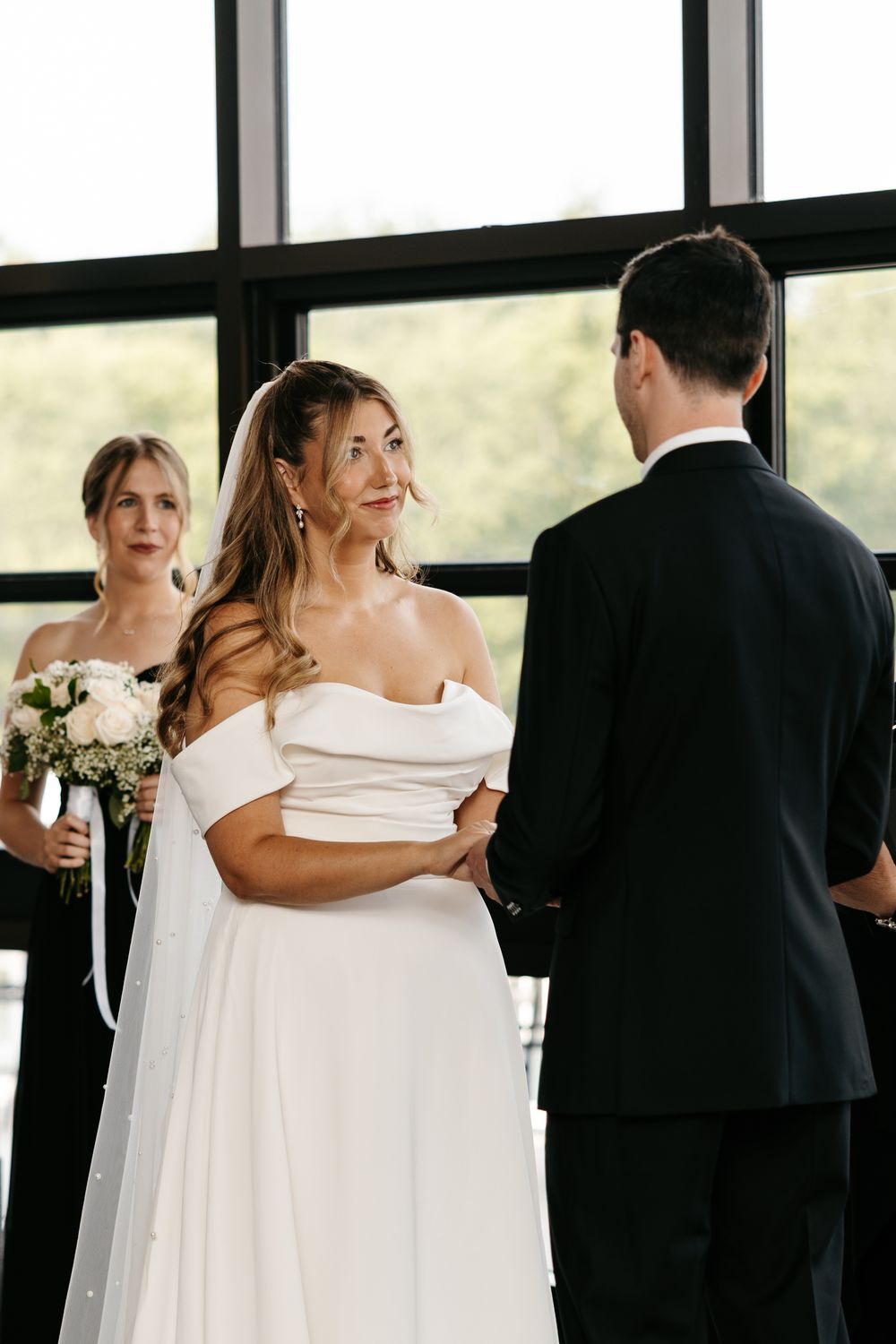 Bride in off-shoulder white gown smiles at groom during indoor wedding ceremony with bridesmaid holding bouquet.