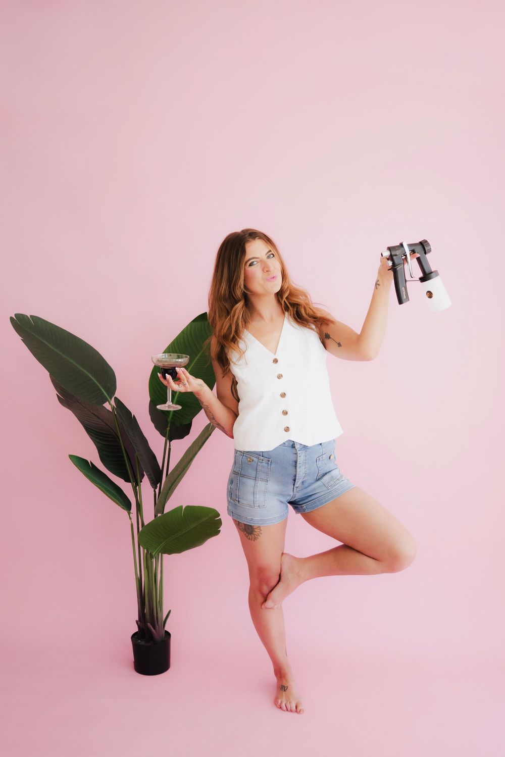 Woman in white top and denim shorts holding camera and wine glass next to tropical plant on pink background.