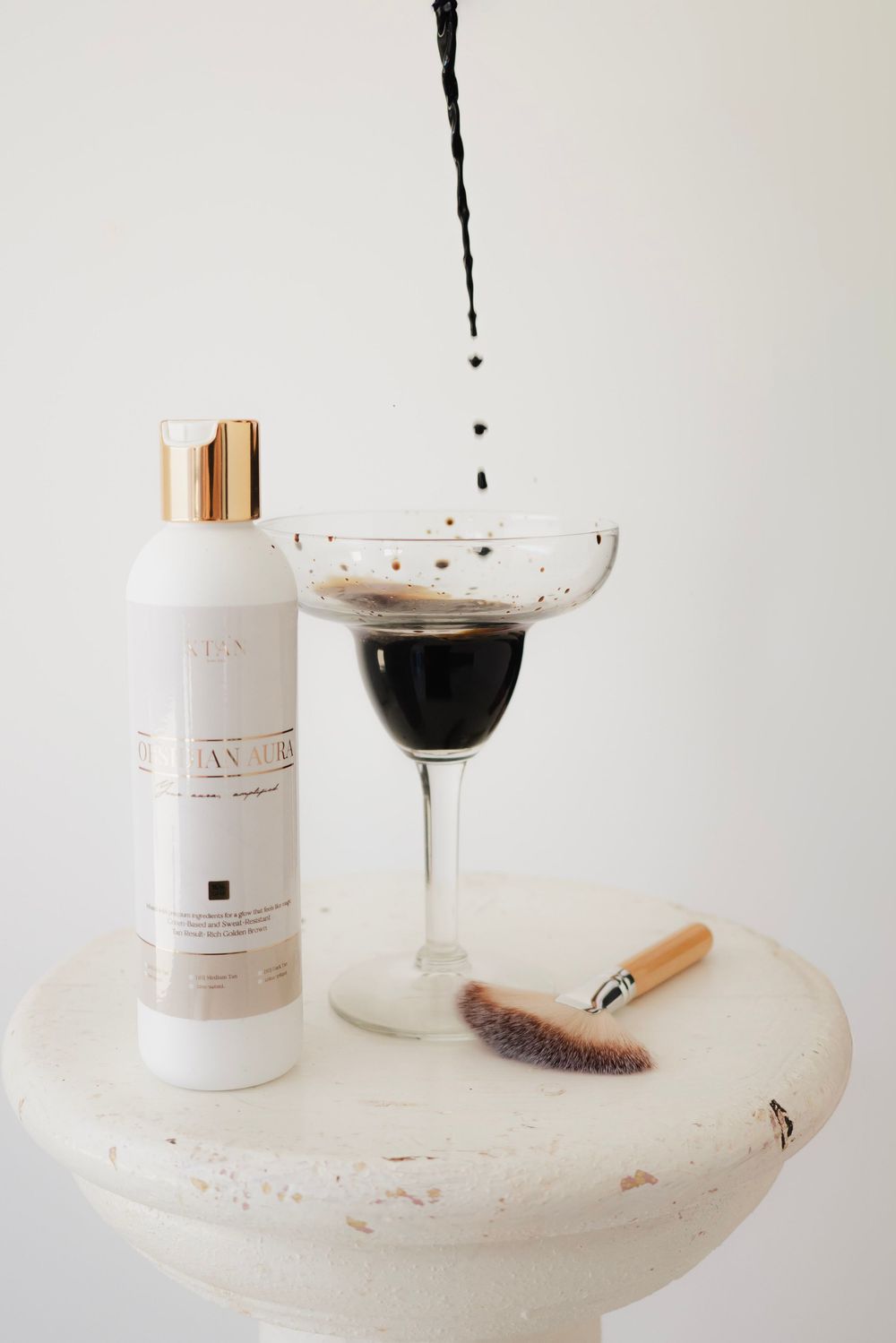 Luxury skincare bottle with gold cap beside a coupe glass filled with dark liquid and a makeup brush on a white pedestal.