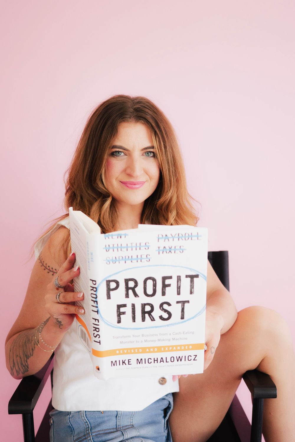Smiling woman holding Profit First book by Mike Michalowicz, sitting in chair against pink background.