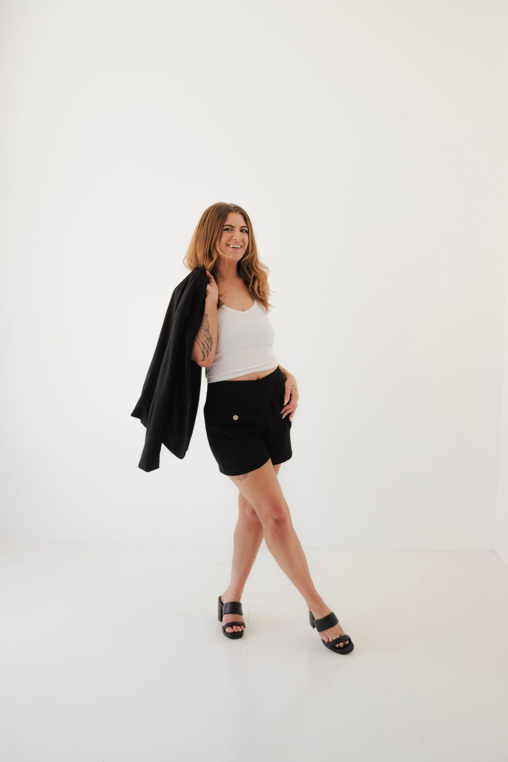 Woman in white crop top, black shorts, and heels, holding a black blazer over her shoulder, smiling in a studio.