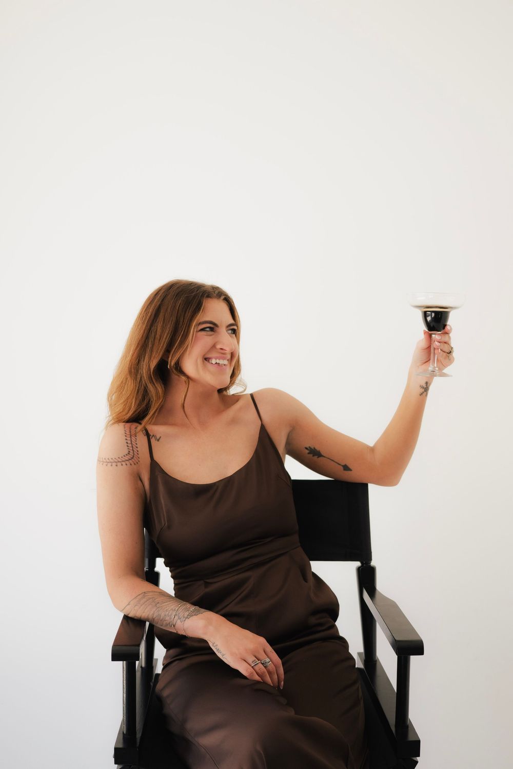 Smiling woman in brown slip dress raising wine glass while seated in director chair.