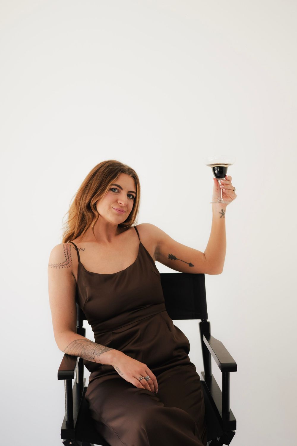 Woman in brown satin dress sitting in director's chair raising a wine glass against white background.