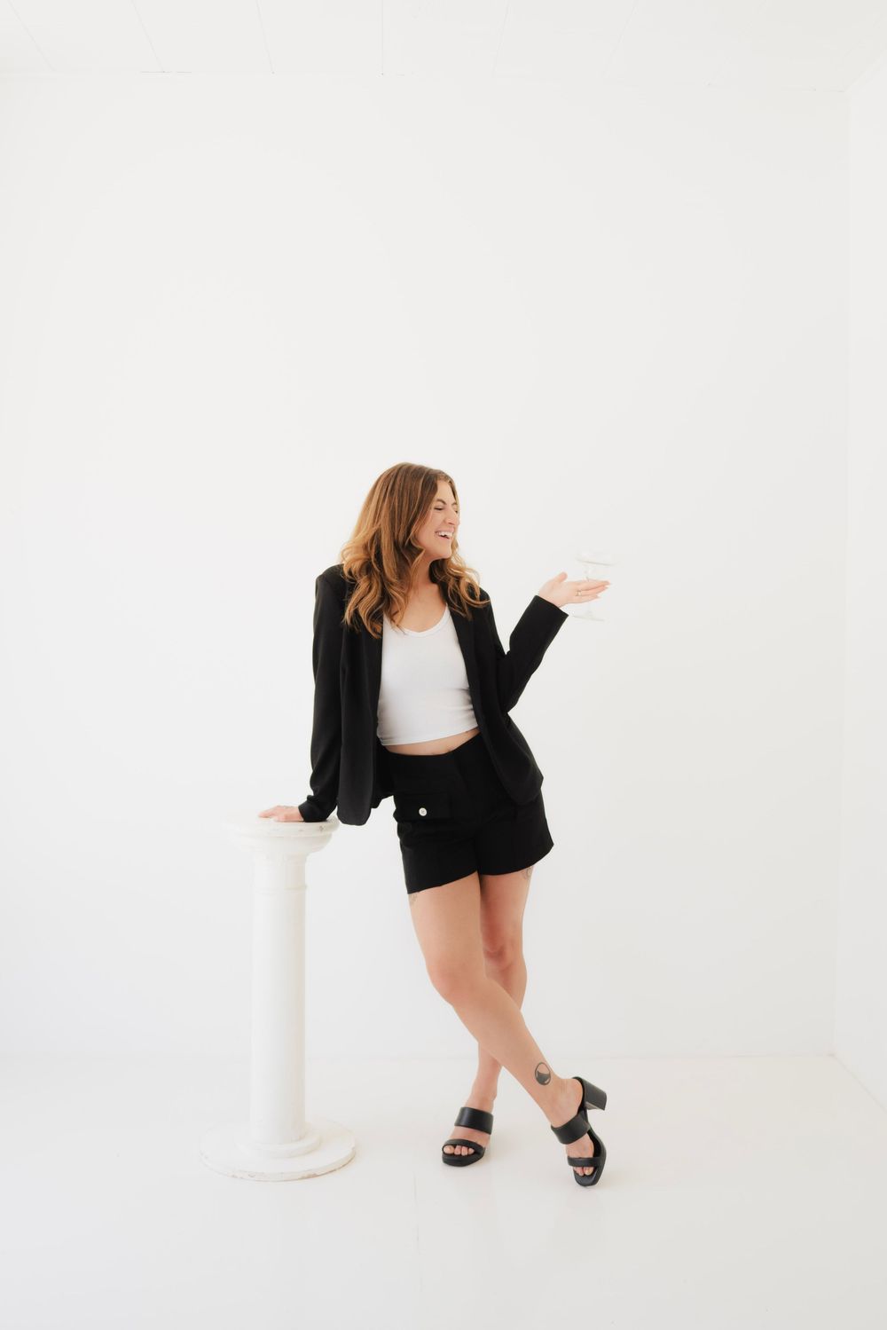 Woman in black blazer and shorts leaning on white pedestal, holding a cup, smiling against white background.