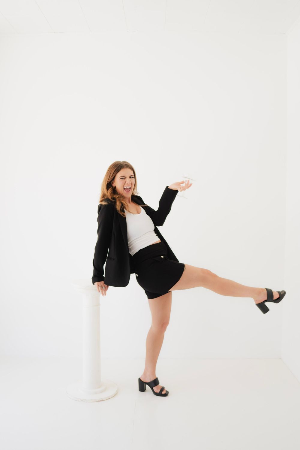 Playful woman in black blazer and mini skirt kicking her leg up beside a white pedestal on a white background.