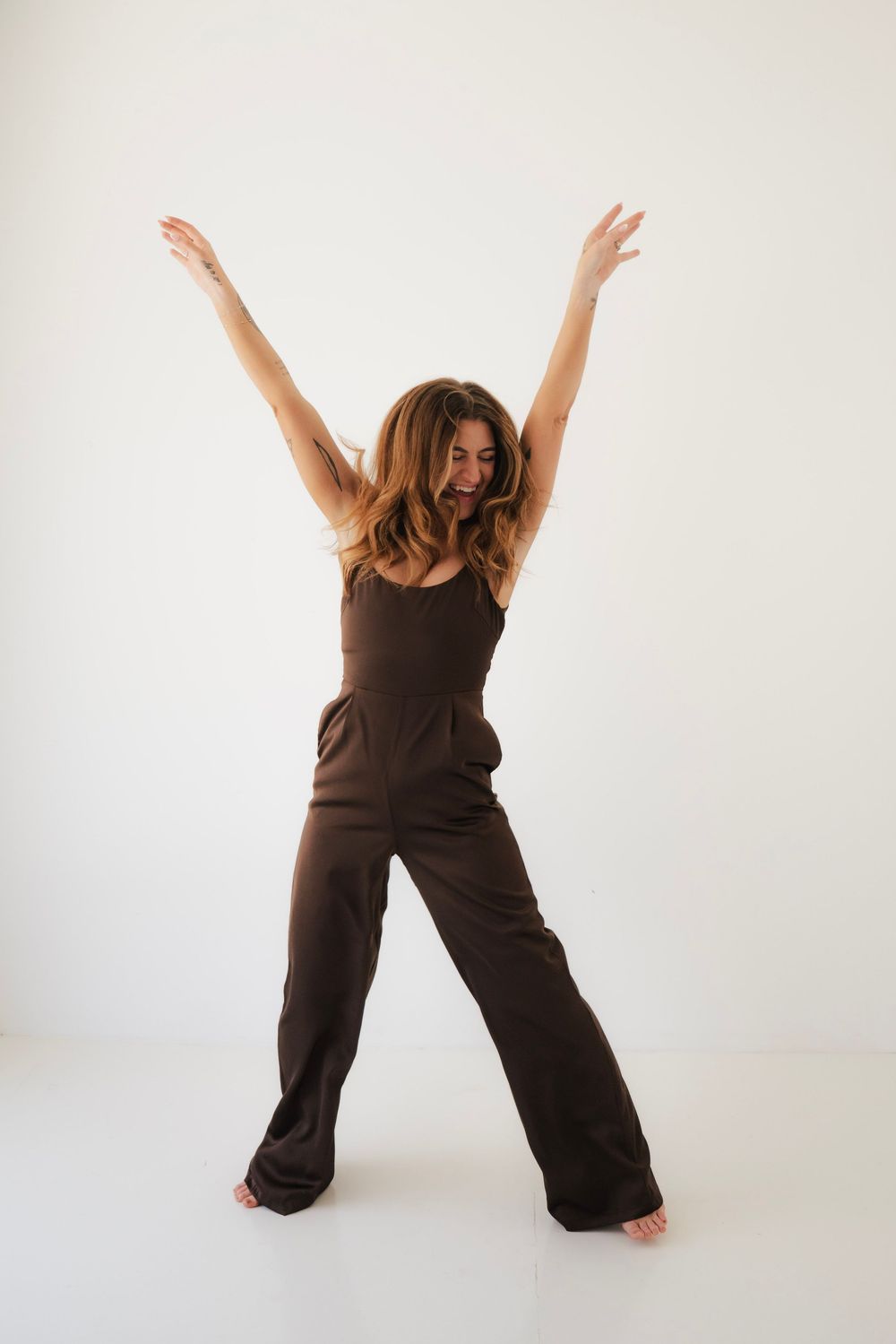 Joyful barefoot woman in brown jumpsuit raising both arms wide open on a white studio background.