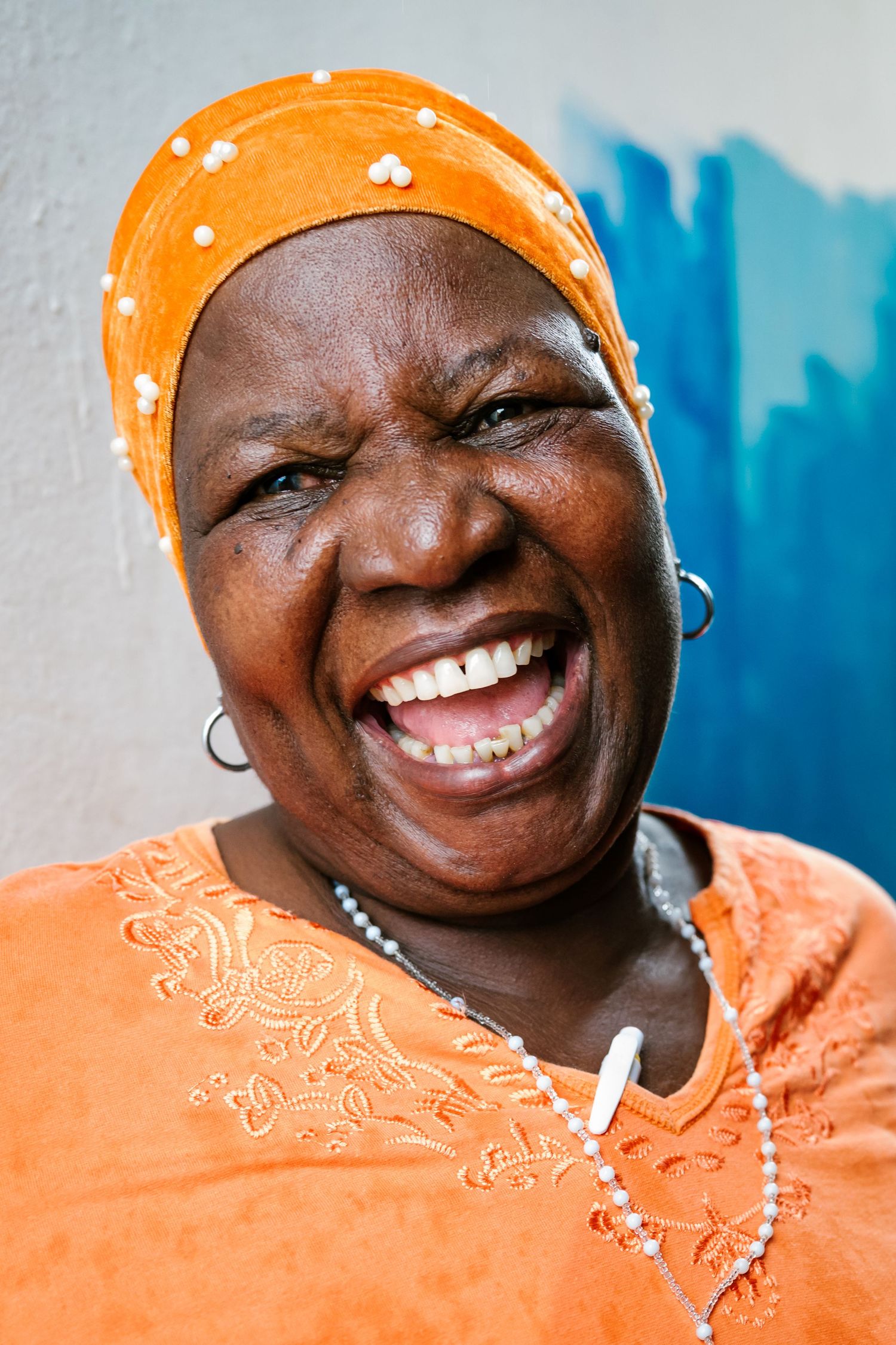 Joyful elderly African woman laughing, wearing orange headwrap and matching top with beaded necklace.