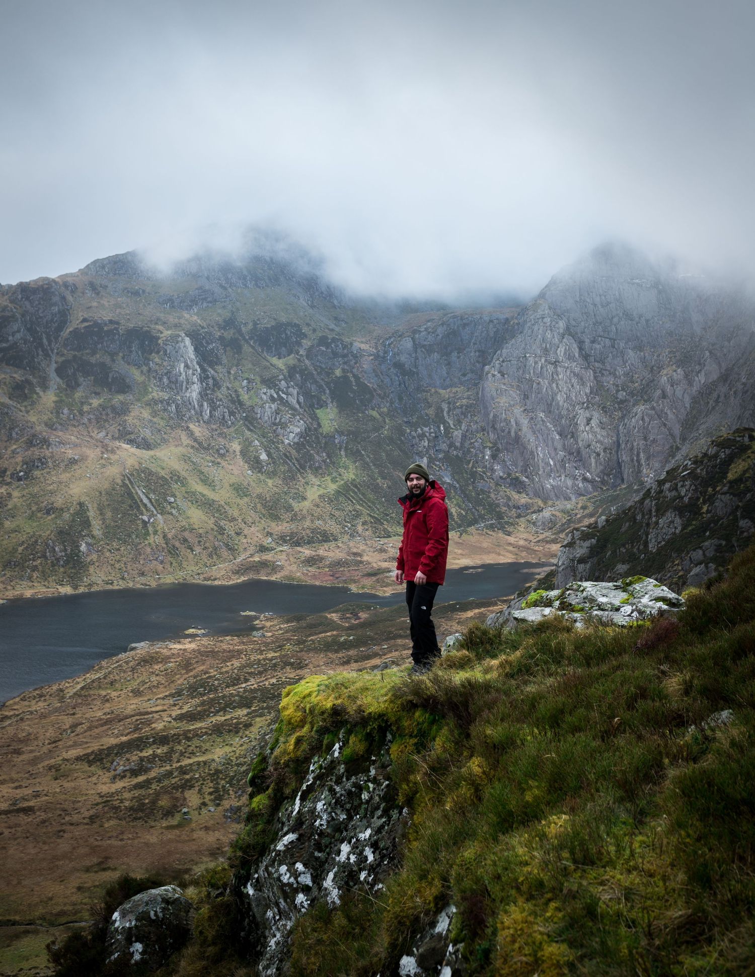 Person in red jacket standing on rocky mountain ridge with misty peaks and alpine lake in background. Snowdonia national park
