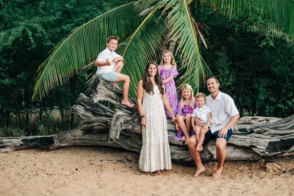 Family of six posing on a fallen log on a tropical beach with palm trees and lush greenery.