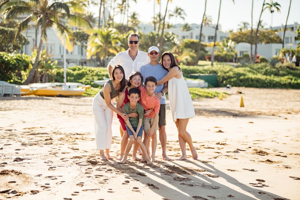 Happy family of seven posing together on a sunny tropical beach with palm trees and resort in background.