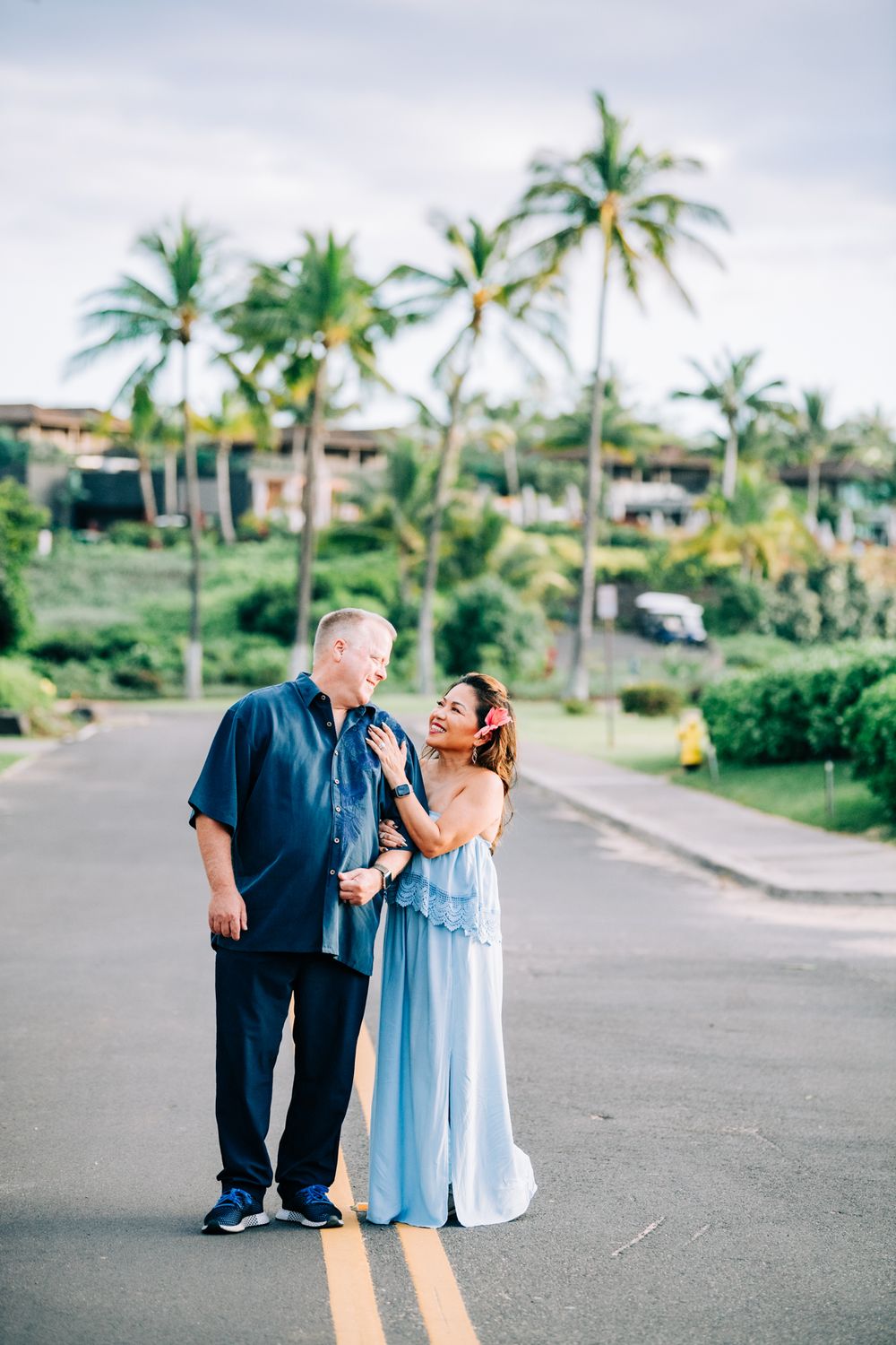 Couple smiling and embracing on a tropical road lined with palm trees, woman in blue gown, man in navy shirt.