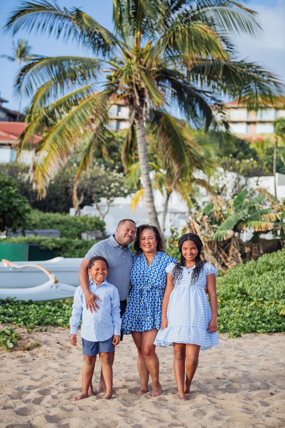 Happy Black family of four posing barefoot on sandy beach with palm tree, wearing coordinated blue outfits on tropical vacation.