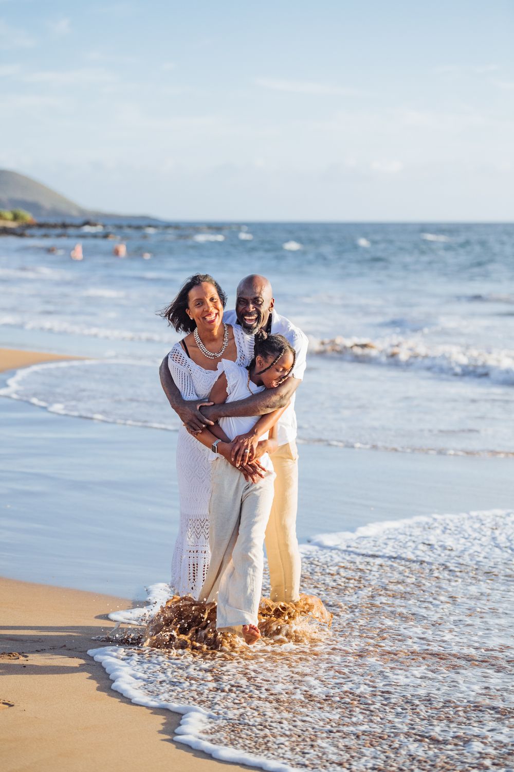 Happy couple embracing on a sunny beach, waves washing over their feet, wearing white and cream outfits.