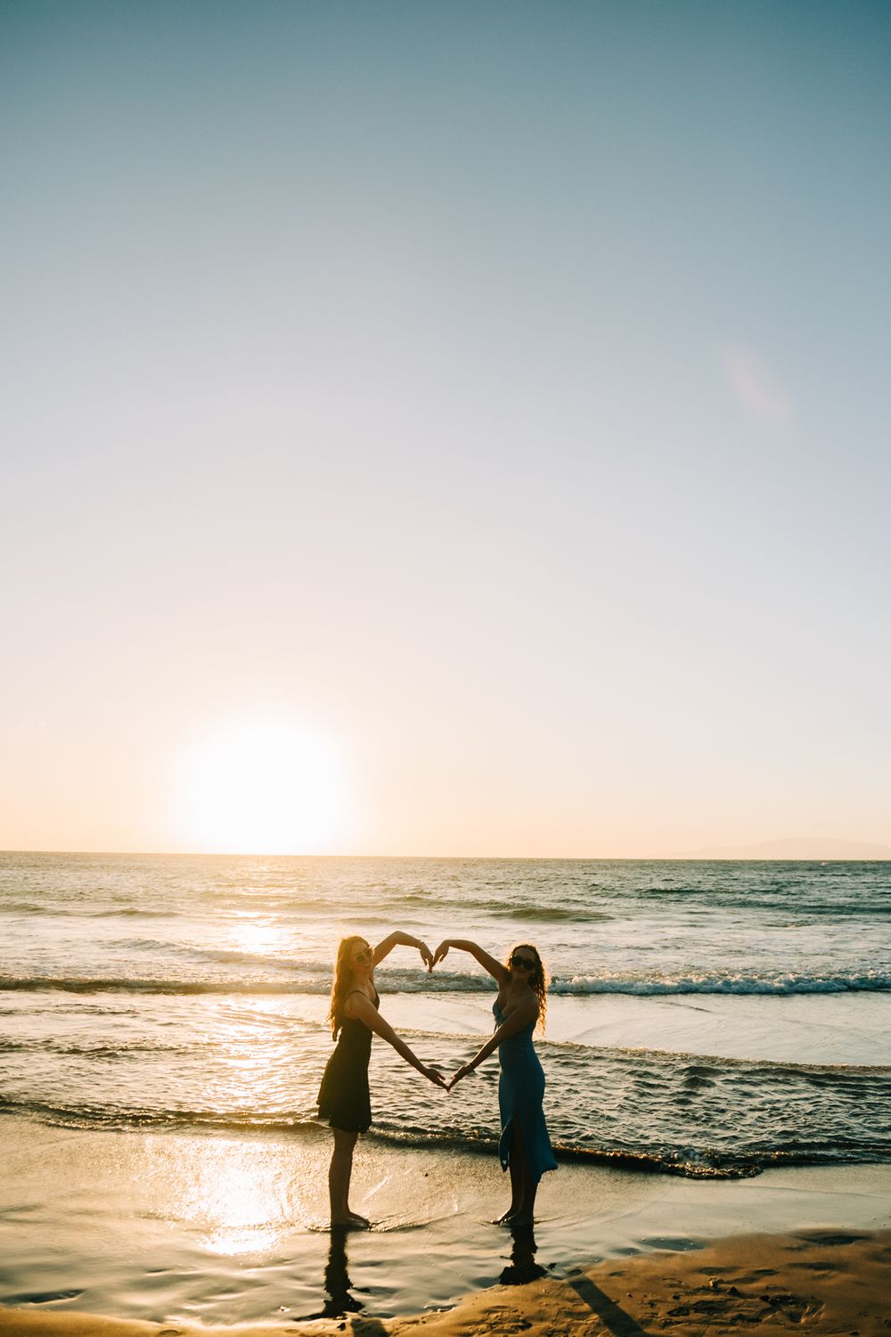 Two women forming a heart shape with their arms at sunset on a beach with golden ocean reflections.