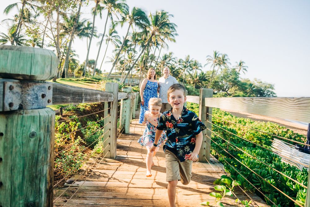Two excited children running along a wooden boardwalk surrounded by tropical palm trees in Hawaii.