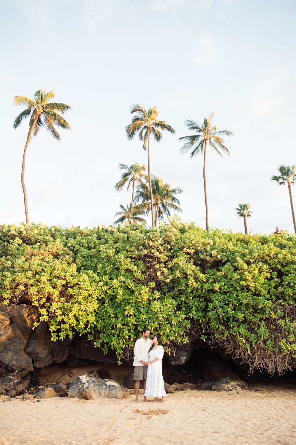 Couple embracing on a sandy beach surrounded by lush tropical greenery and tall palm trees.