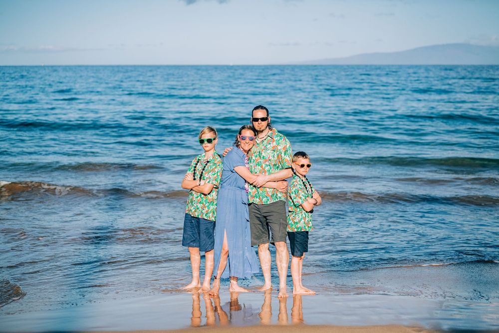 Family of four in matching Hawaiian shirts posing on a sunny beach with ocean waves behind them.