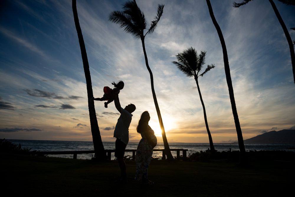 Silhouette of family at sunset with palm trees on tropical beach, parent lifting child in air.