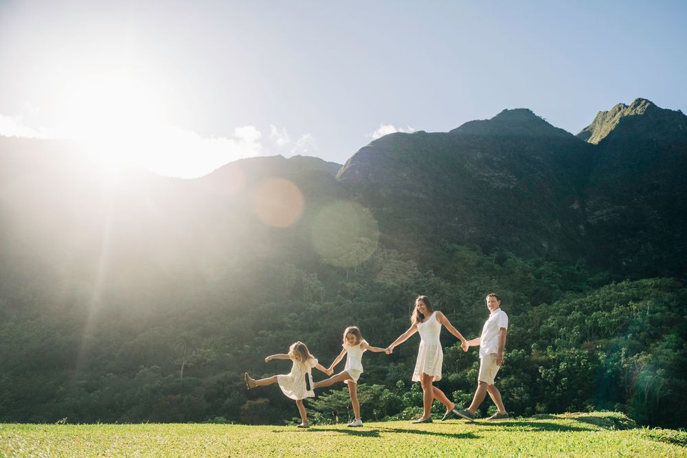 Family in white outfits holding hands walking on green grass with stunning mountain backdrop in bright sunlight.