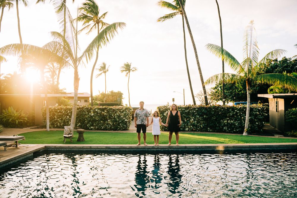 Family of three standing by a resort pool at golden sunset with tall palm trees in Hawaii.