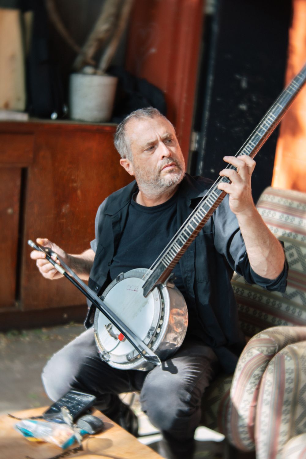 A bearded man plays a banjo in a rustic room filled with vintage furniture.