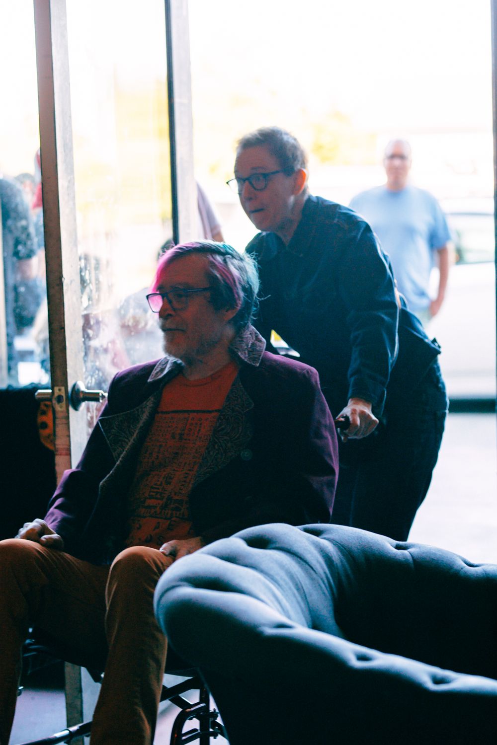 Person in wheelchair with pink hair and purple jacket, assisted by another individual near a glass door indoors.