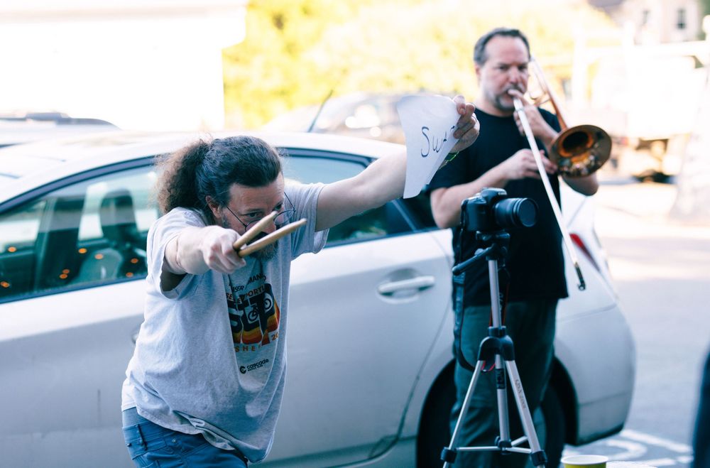 Musician plays trombone outdoors near a car while another person poses with drumsticks beside a camera tripod.