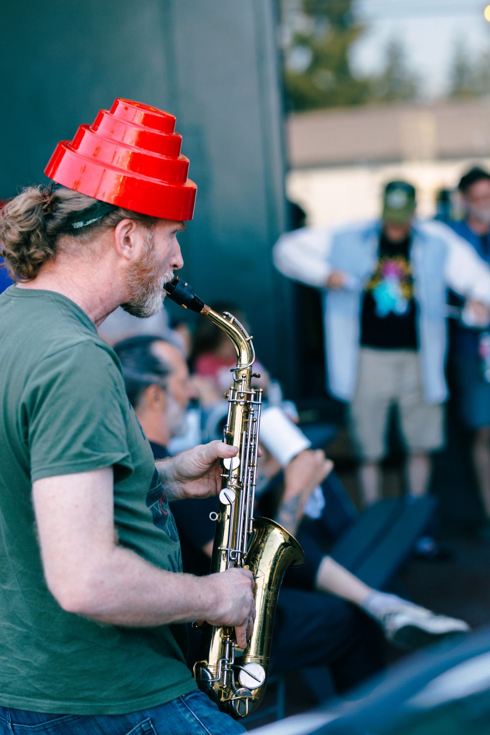 Man in red novelty hat plays saxophone at outdoor festival event.